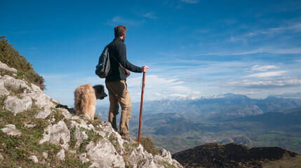 Hombre joven barbudo  con palo de madera y perro en montaña de Asturias mirando el horizonte