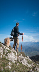 Hombre joven barbudo  con palo de madera y perro en montaña de Asturias mirando el horizonte