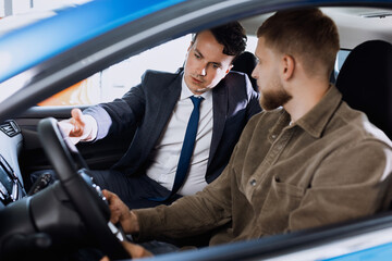 An employee of a car dealership shows new car options to a male buyer sitting in the salon