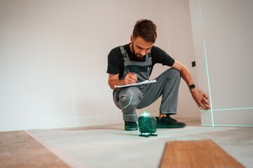 Making notes into the document and using laser leveler. Man is installing new laminated wooden floor
