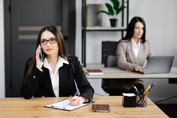 Attractive young business woman wearing jacket talking on mobile phone while sitting on a desk and using notebook near her colleague at work in office