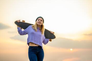 Young woman with skateboard walking smiling, sunset background, trendy teenager of generation z
