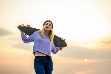 Fotobehang Young woman with skateboard walking smiling, sunset background, trendy teenager of generation z © MandriaPix
