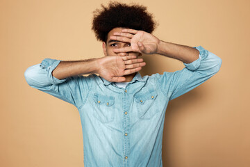 Portrait of young latin curious silent male with hands covering mouth and one eye looking aside peeping, while being scared of something, isolated on brown studio background in blue denim shirt