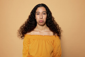 Portrait of adorable offended sad young female of 20s with dark skin and hair looking at camera with upset frustrated facial expression pursing lower lip and grimacing sadly on brown background