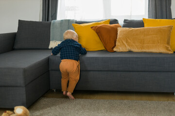 Toddler boy laughing having fun standing near sofa in living room at home copy space. Adorable baby making first steps alone. Happy childhood and child care concept