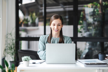 Young confident businesswoman sitting at the office table working on laptop computer. Business concept