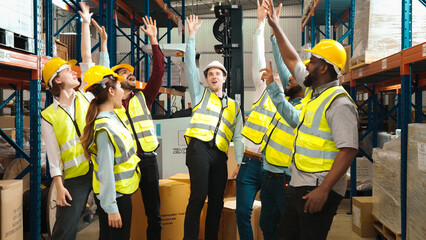 Multiracial warehouse workers in safety wear are stacking hands and cheering to celebrate after finishing a job in warehouse. Male and female logistic employees are enjoying the achievement together.