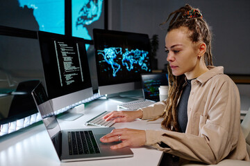 Young serious female programmer looking at data on laptop screen while sitting by workplace in dark office and working overtime