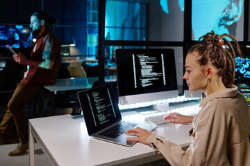 Serious female programmer with dreadlocks decoding data on laptop screen while sitting by desk with...