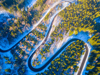Asphalt road serpentine in snowy wintertime. Cold winter and sunny day above forest road with illuminated trees by rising sun. Aerial view from drone.