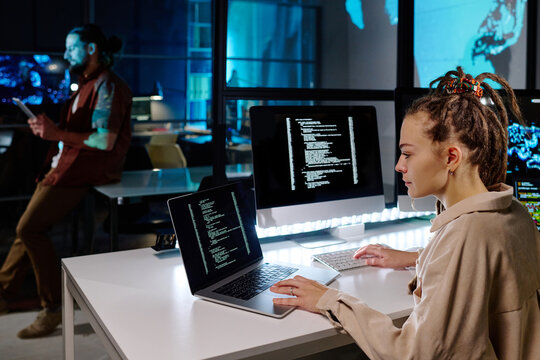 Serious female programmer with dreadlocks decoding data on laptop screen while sitting by desk with computer monitor in dark office