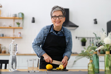 Smiling mature woman cuts a yellow lemon for a fruit drink at home in the kitchen.