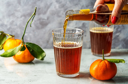 Chinotto refreshing citrus drink in a glass, woman's hand holding bottle and pouring drink to the glass, light grey surface with grey concrete background