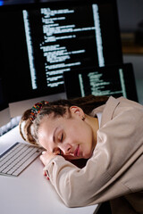 Young tired woman with dreadlocks keeping head on desk by keyboard while sleeping in dark office against computer screens with coded data