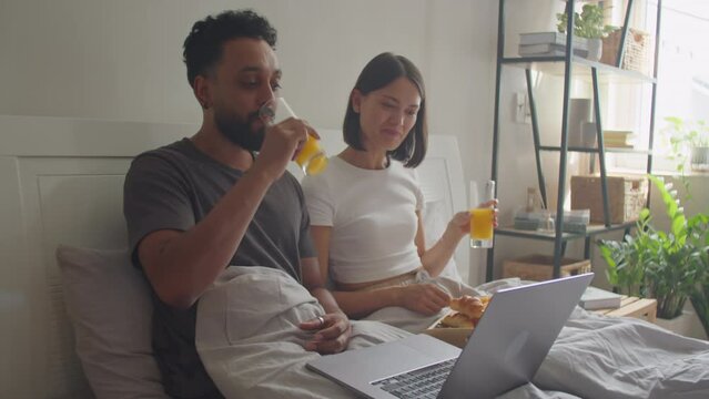 Young married couple having breakfast in bed, watching movie on laptop and smiling while spending time together in morning at home