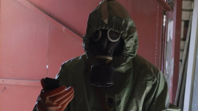 Man wearing green respirator and mask checks level of radiation in small garage. Professional specialist measures radiation indicators with device