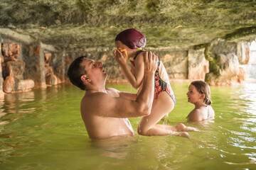 Fototapeta premium Family, dad and children bathe, swim in outdoor dirty hot springs. Thermal water bath pool and healthy natural, clay and yellow green mud. Spa wellness relax famaly winter vacation in mountain hotel