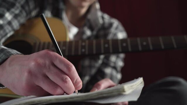 young man writing song lyrics with acoustic guitar in cozy room.