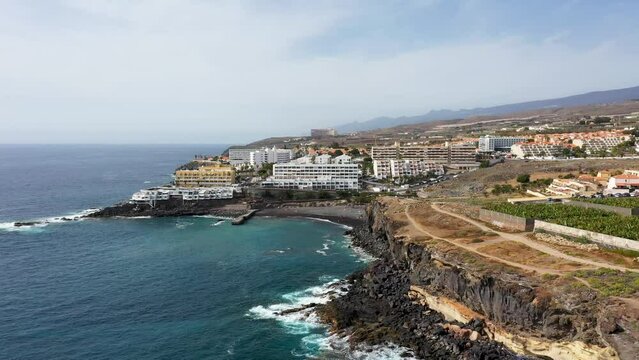 Aerial drone footage of the beautiful town of Costa Adeje, Santa Cruz de Tenerife in Spain showing the Playa de Ajabo beach with it's black sand, and hotels and businesses in the town