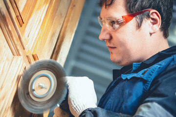 Man treats wooden surface. Worker in safety glasses grinds wooden door with metal brush....