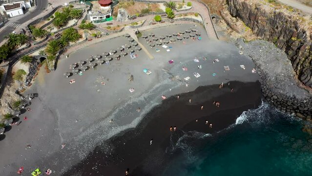 Aerial drone footage of the beautiful town of Costa Adeje, Santa Cruz de Tenerife in Spain showing the Playa de Ajabo beach with people relaxing and having fun on the black sandy beach.