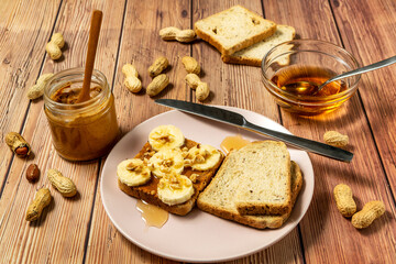 Peanut butter or peanut paste in an open jar and spread on a toast sandwich with banana and honey and peanuts in shells scattered on the table