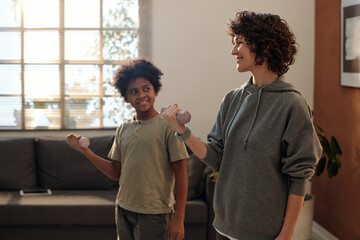 Happy young woman and her adorable son in activewear practicing exercises with dumbbells at home while standing in front of camera