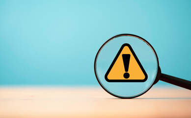Yellow triangle caution warning sign inside of magnifier glass on blue background and copy space for maintenance notification error and risk concept.