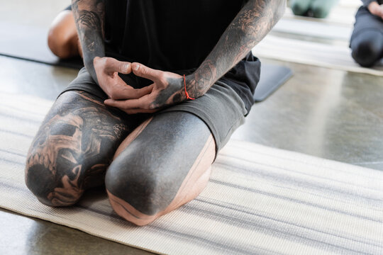Cropped view of tattooed man sitting in Thunderbolt asana on yoga mat in studio.