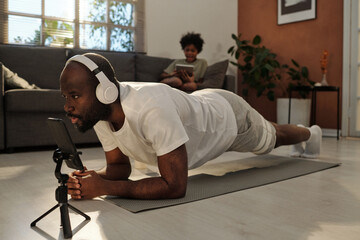 Young man in headphones and activewear repeating plank after fitness trainer on smartphone screen while exercising on mat at home