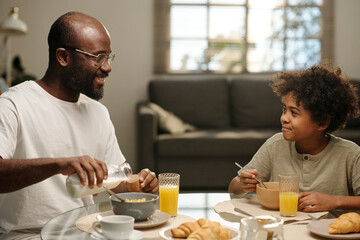 Fototapeta na wymiar Young African American man in eyeglasses pouring milk in bowl with muesli and talking to his cute son while having breakfast with him