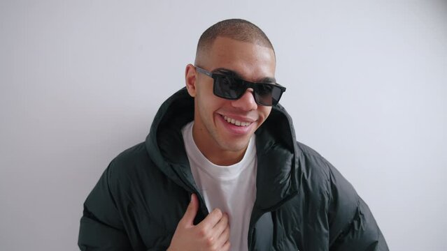 Studio shot of confident street style loving man in sunglasses, putting on his black puffer jacket against white background. High quality 4k footage