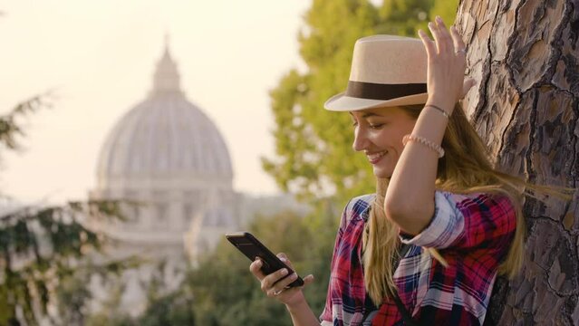 Happy young blonde woman with long hair and blue eyes laining on a tree, using her smartphone in front of Rome cityscape. Saint Peter church from Gianicolo panoramic view.