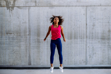 Fototapeta premium Young multiracial girl jogging and jumping in city, in front of concrete wall, side view.