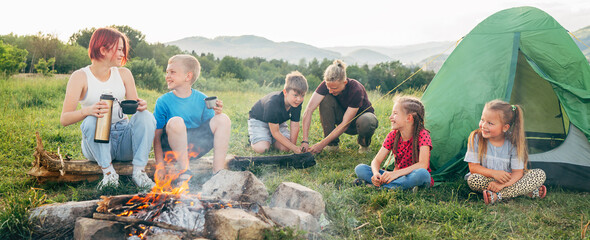 Group of smiling kids has a merry conversation near a smoky campfire. They drinking tea from a...