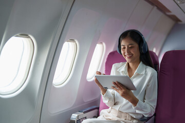 Young Asian attractive business woman passenger sitting on business class luxury plane while wearing wireless headphone for relax and working with tablet during the flight