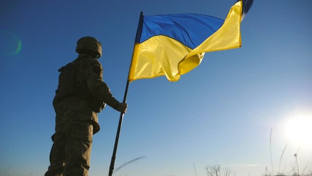 Male soldier of ukrainian army lifting national banner against blue sky. Man in military uniform holds a waving flag of Ukraine. Victory against russian aggression. Invasion resistance. Low view