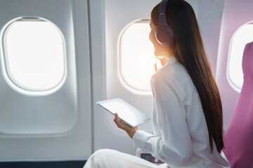 Young Asian attractive business woman passenger sitting on business class luxury plane while wearing wireless headphone for relax and working with tablet during the flight