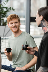 positive man with red hair holding Japanese cup with puer tea near tattooed friend in yoga studio.