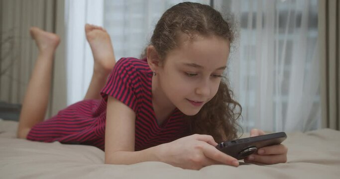 Phone for gaming. Child playing video gameat home lying on his bed. Beautiful child girl 10 years old playing mobile game on smartphone at home. Adorable kid girl playing mobile phone lying on bed. 4K