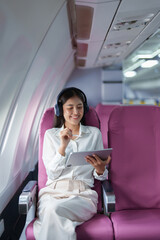 Young Asian attractive business woman passenger sitting on business class luxury plane while wearing wireless headphone for relax and working with tablet during the flight