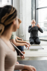 Mature coach practicing nostril breathing near blurred interracial group in yoga class.