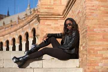 young and beautiful black latin woman wearing black clothes and sunglasses is sitting on the steps of a square in seville, spain. The photo is taken in profile and the girl is doing different poses.