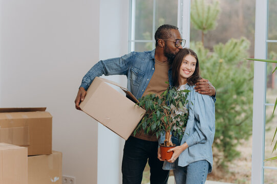 A happy young family is moving into a house. A black man and woman are holding cardboard boxes for moving, smiling.