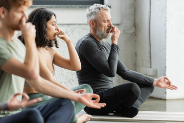 Mature man practicing nostril breathing and gyan mudra near interracial people in yoga studio.