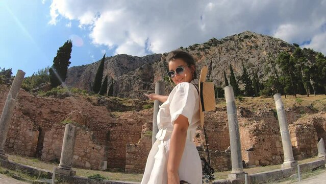 Young woman visiting ruins of temple of Apollo in Delphi, archaeological site by mount Parnassus, Greece, Europe. Large hat, fashion white dress.
