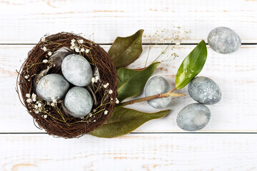 Brown nest of twigs with three gray Easter eggs, feathers, dried magnolia leaves and sprigs of gypsophila on wooden background. Minimalistic Easter concept