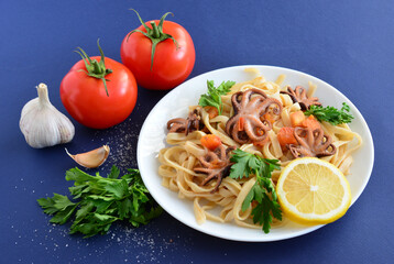 A plate of pasta with baby octopus, a lemon and tomatoes on the side
