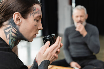 side view of tattooed man holding Chinese cup with brewed puer tea near middle aged man on blurred background.